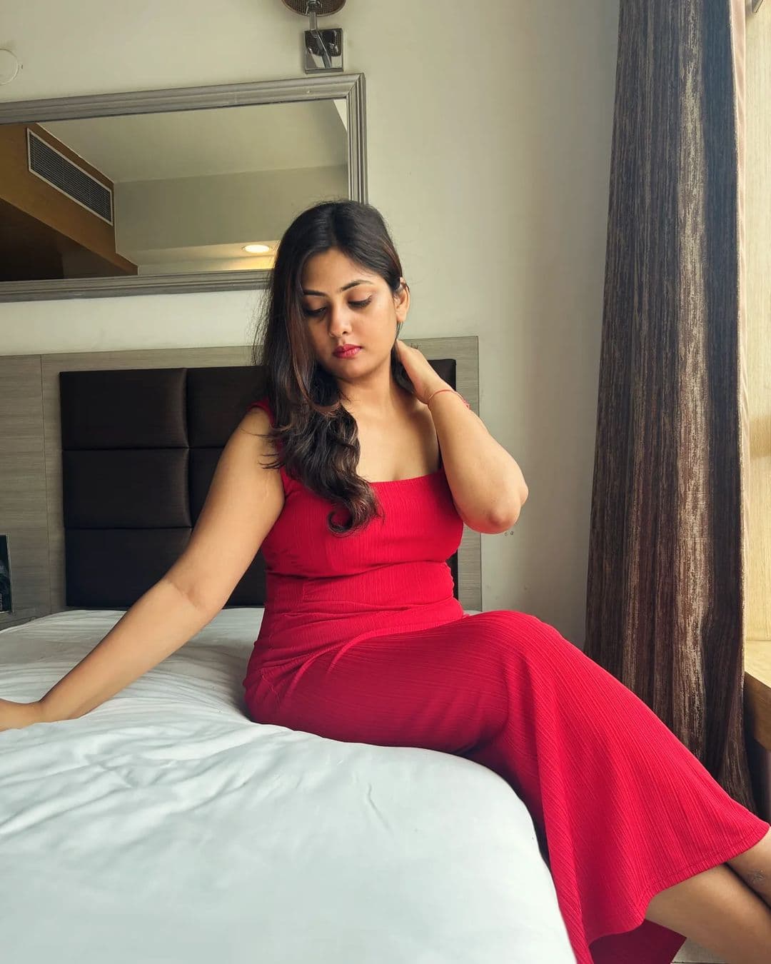 Woman in a red dress sits on a bed, looking down while touching her neck.